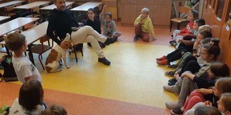 Powiększ grafikę: Dzieci siedzą na podłodze i słuchają pana Marcina, przy którym stoi pies Latek.