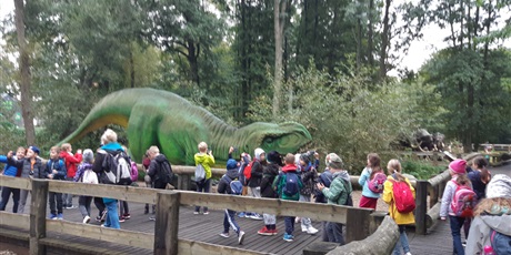 Powiększ grafikę: Dzieci idą po ścieżce edukacyjnej i oglądają dinozaury.